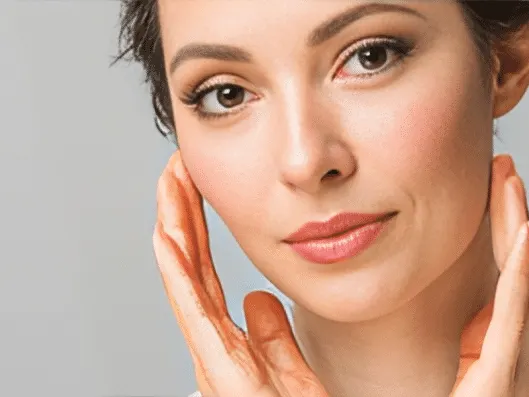 A woman with smooth skin cupping her jawline with both hands against a gray background