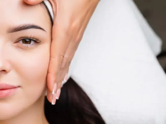 A woman lying on a white towel while hands gently hold both sides of her face during a facial treatment