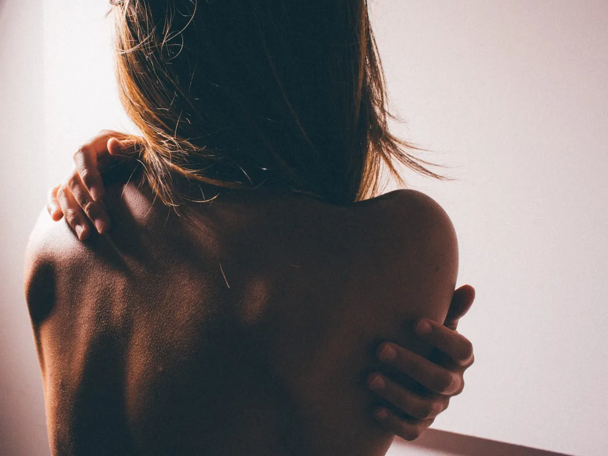 A woman seen from behind, holding her bare shoulder with one hand
