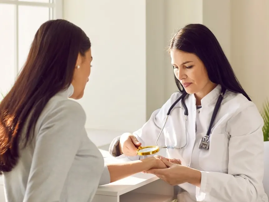 A dermatologist in a white coat examining a woman's hand with a magnifier at a clinic desk