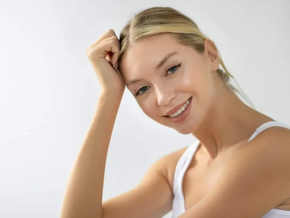 A smiling blonde woman in a white top leaning her head on her fist against a light background