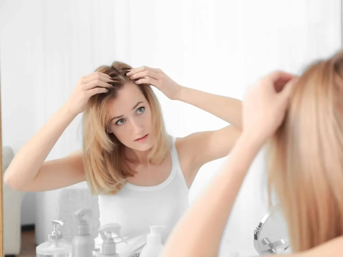 Woman in her 30s looking at hair and scalp in mirror, experiencing hair loss