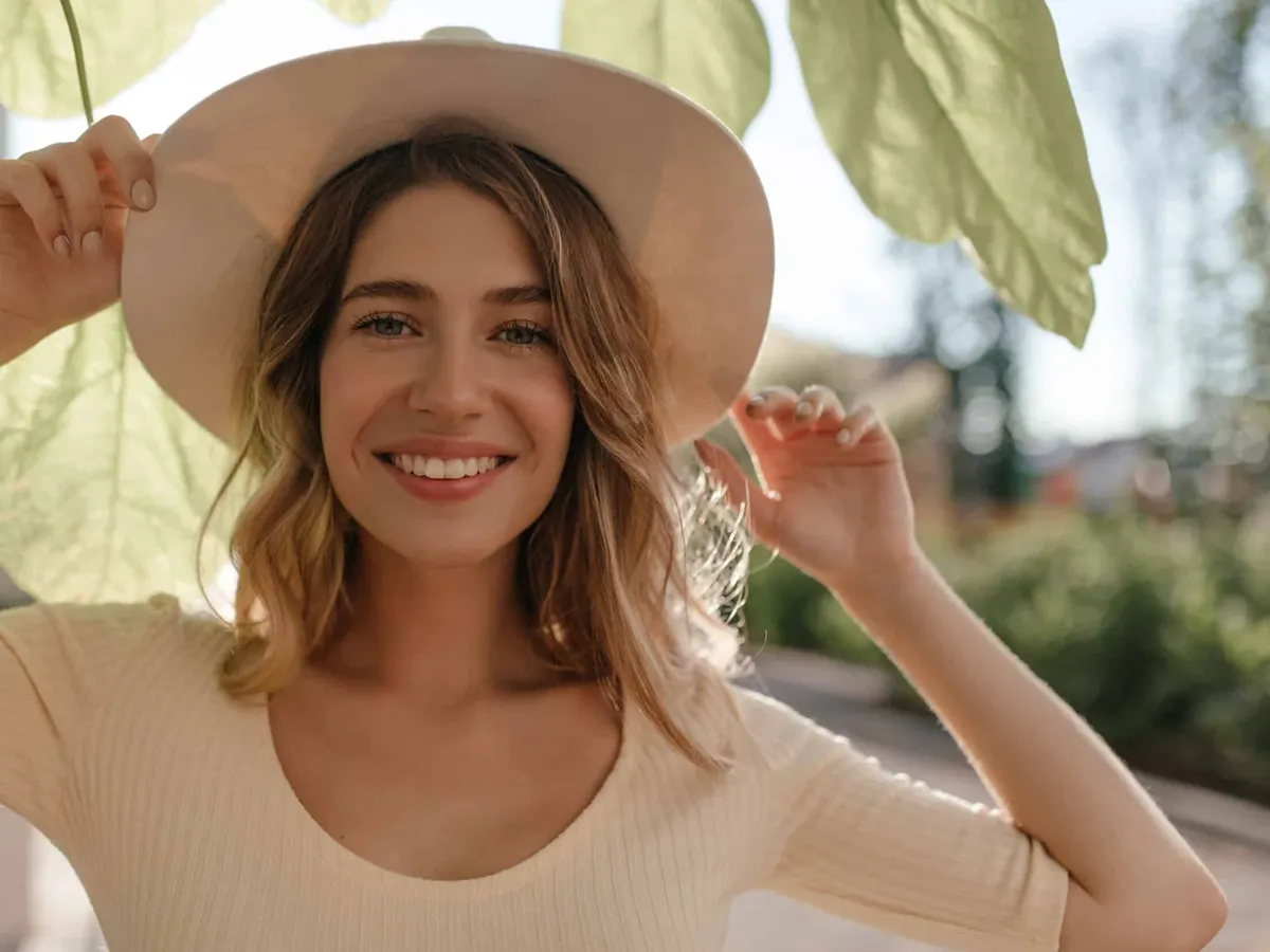 woman smiling with teeth in stylish hat, hiding from sun on light background close-up