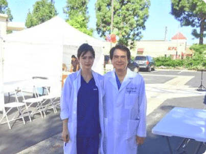 Two clinicians in white coats standing beside a white event tent in a parking lot