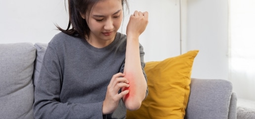 A woman on a sofa scratching a red, irritated patch on the inside of her forearm