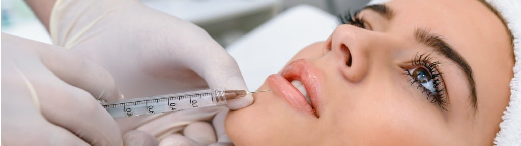 A woman lying back as a gloved clinician injects near her upper lip with a syringe