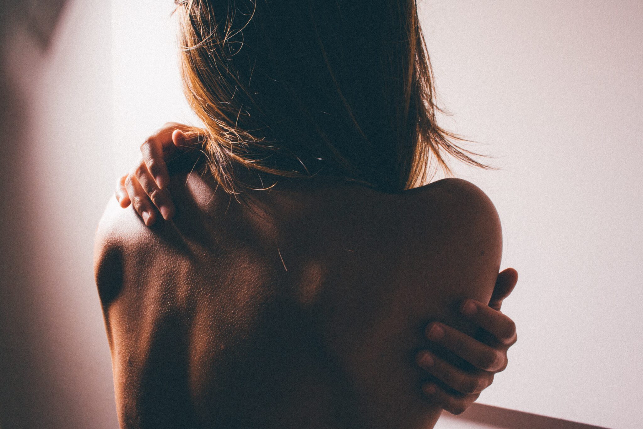 A woman seen from behind, holding her bare shoulder with one hand