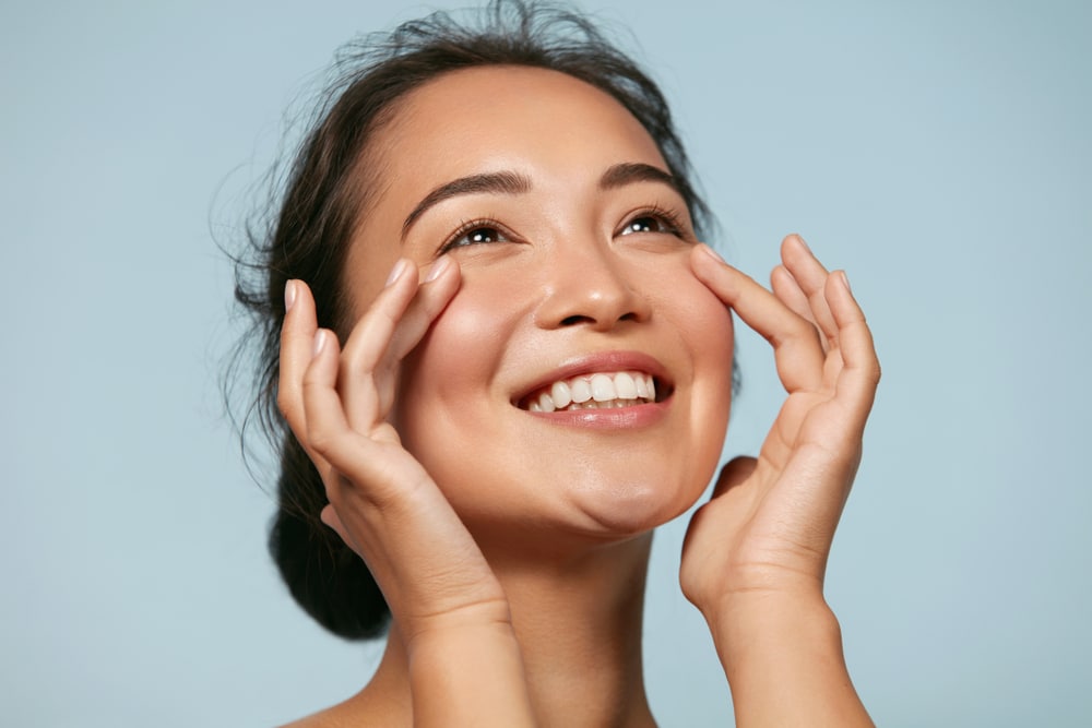 A smiling woman with dark hair touching her cheeks with her fingertips against a blue background
