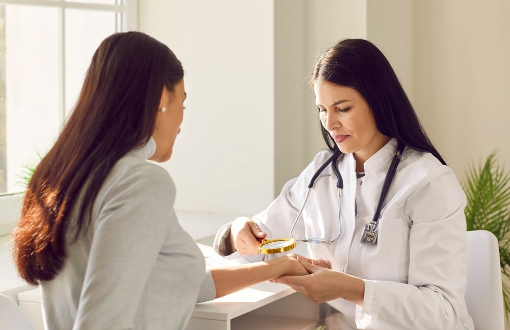 A dermatologist in a white coat examining a woman's hand with a magnifier at a clinic desk