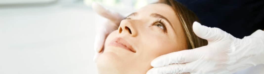 A woman with her face tilted up as a clinician in white gloves holds her face during a skin treatment