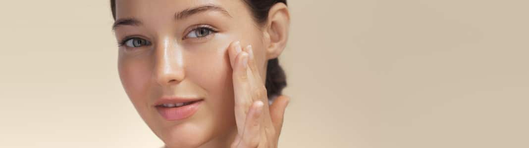A woman with glowing skin touching her cheek with her fingertips against a soft beige background