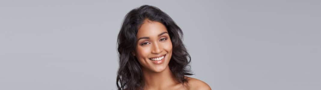 A smiling woman with long dark wavy hair and bare shoulders against a gray background