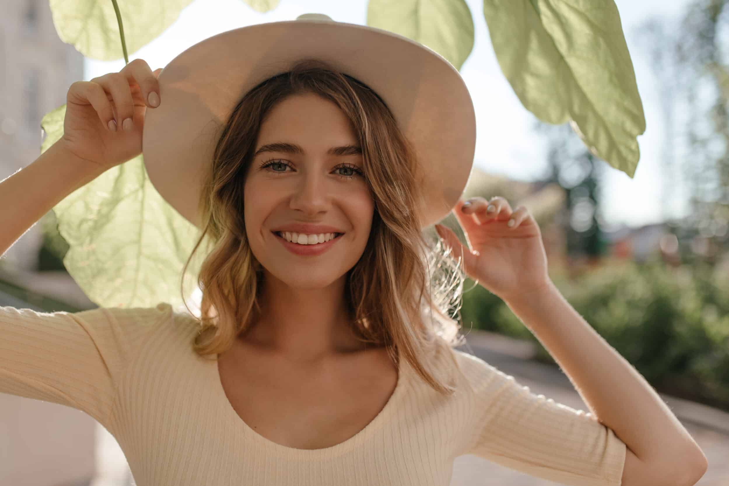 woman smiling with teeth in stylish hat, hiding from sun on light background close-up